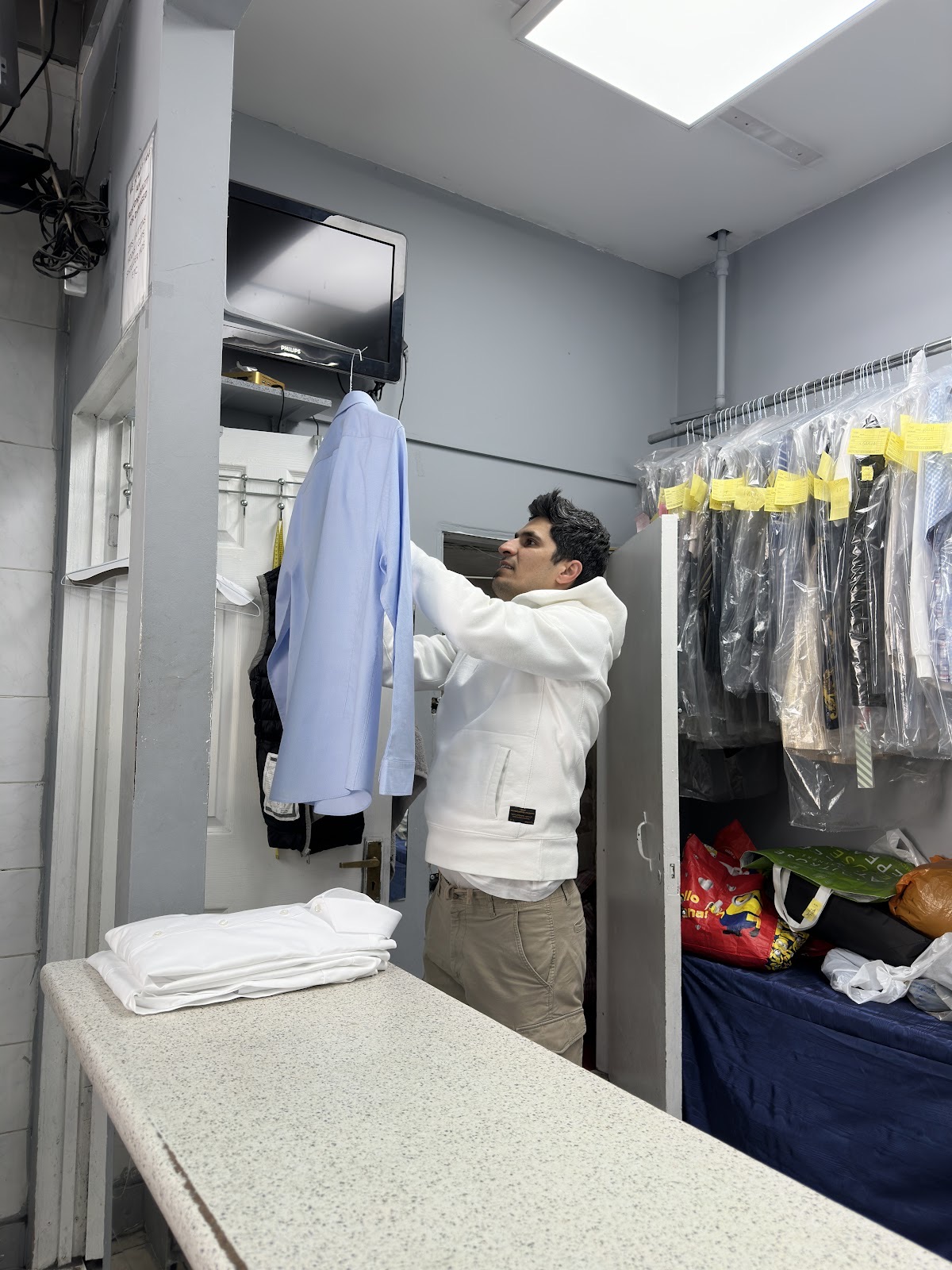 Inside the shop, a freshly cleaned blue shirt being hung on the rail beside dry-cleaned garments in clear covers, folded white linen stacked on the counter
